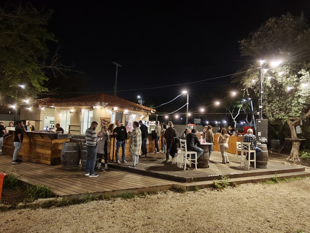 תמונת לילה של הפאב החברתי במזכרת בתיה. בבר עץ מואר באור חם עומדים אנשים רבים ומפטפטים. מעל הבר תלויות שרשראות נורות. האווירה נעימה וקהילתית.